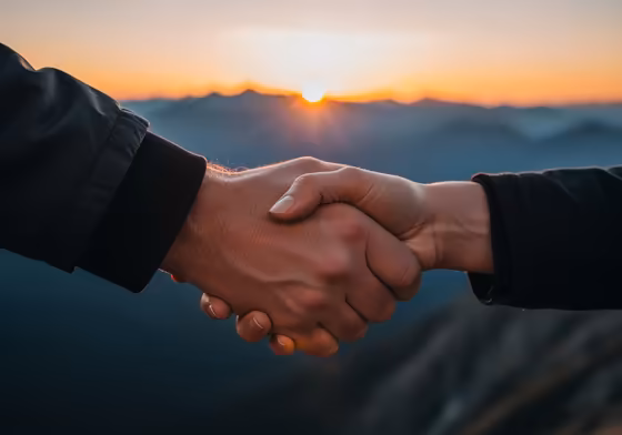 Two hands clasped together, symbolizing partnership, respect, and strength, with a rugged, natural landscape in the background during sunset.